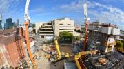 "Yellow crane lifts pallets of construction material. Heritage building to the left, core of lifts being constructed to the right."